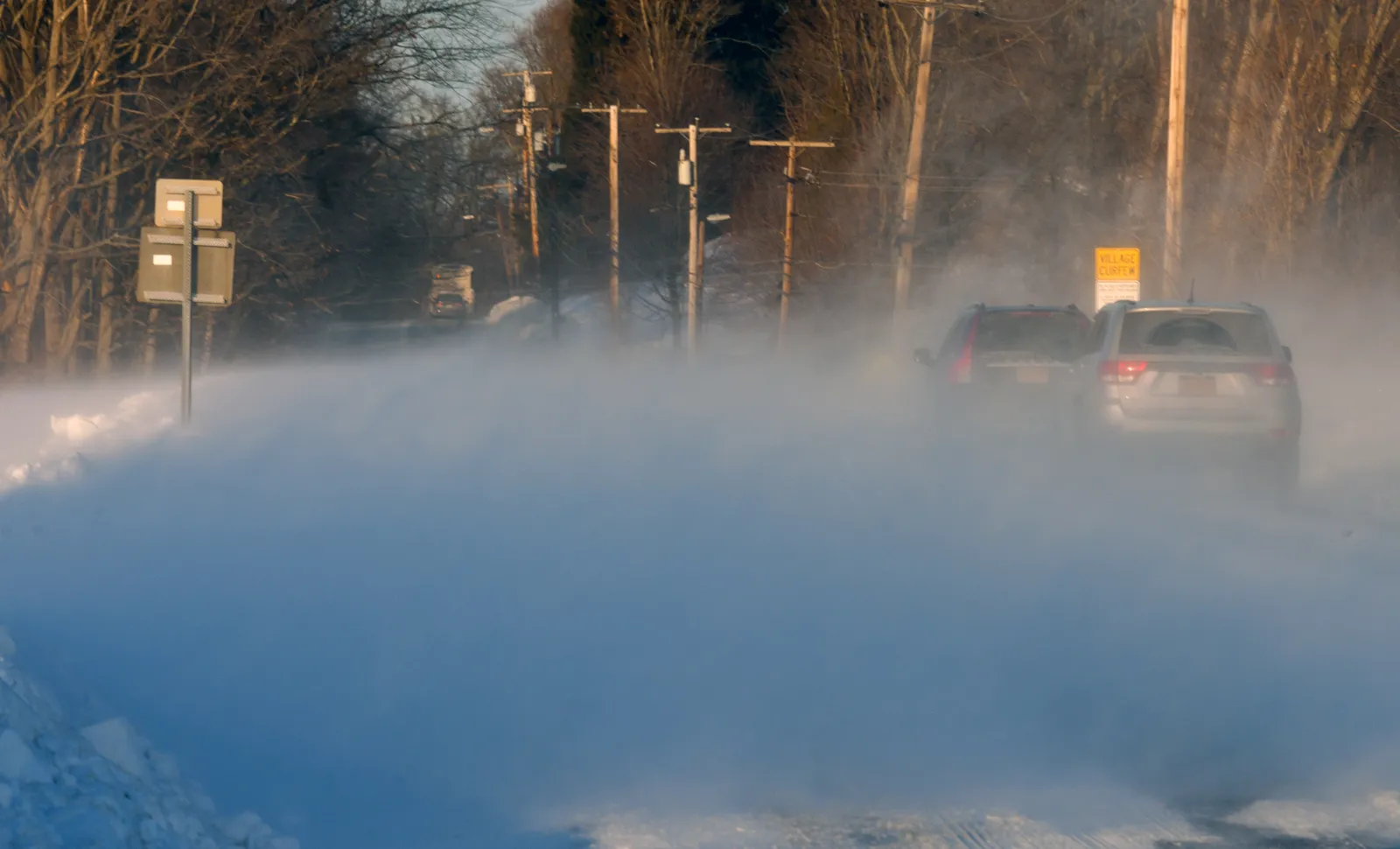 Two cars crawling through blowing snow on a rural highway in low winter sun